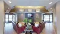 This single-floor space presents a well-appointed communal lounge area designed with both comfort and style in mind. The room is anchored by a striking floor-to-ceiling stacked stone fireplace, above which a wall-mounted flat-screen television is centered as a focal point. Three large drum-style pendant lights with warm-toned shades hang from a coffered ceiling, casting an inviting glow throughout the space and adding a refined architectural detail overhead.

The seating arrangement is thoughtfully composed, featuring rich burgundy leather armchairs and a coordinating sofa positioned around a dark rectangular coffee table. Decorative accents, including a glass vase with tropical greenery and a lantern centerpiece, add a fresh, organic touch to the tableau. A patterned area rug grounds the seating group atop light hardwood flooring that extends throughout the room.

The walls are finished in a soft gray tone, complemented by tasteful framed wall art and decorative panels flanking the fireplace. Natural light filters in through glass-paneled doors visible on either side of the space, enhancing the open and airy feel. The overall ambiance is polished and welcoming, making this lounge well-suited for relaxing or socializing in a refined communal setting.