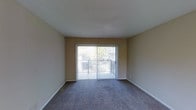 This single-floor residential space offers a straightforward and functional layout designed for comfortable everyday living. The main living area is a standout feature, characterized by wall-to-wall carpeting in a neutral gray tone that adds warmth underfoot. Smooth, light-colored walls and a clean white ceiling create a bright and open atmosphere throughout the room.

A large sliding glass door serves as the focal point of the living area, allowing an abundance of natural light to flood the interior. Vertical blinds provide adjustable light control and privacy. The sliding door opens directly onto an outdoor balcony enclosed by a metal railing, offering a pleasant outdoor extension of the living space with views of surrounding greenery and neighboring structures.

The overall design is simple and understated, with neutral beige tones on the walls complementing the carpeted floors. White baseboards provide a crisp contrast and a finishing detail throughout. The space is vacant, making it easy to envision a variety of furniture arrangements and interior styles. The single-floor layout ensures ease of accessibility and movement throughout the home. This space is well-suited for those seeking a low-maintenance, functional living environment with the added benefit of natural light and outdoor access.