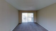 This single-floor residential space offers a straightforward and functional layout designed for comfortable everyday living. The main living area is a standout feature, characterized by wall-to-wall carpeting in a neutral gray tone that adds warmth underfoot. Smooth, light-colored walls and a clean white ceiling create a bright and open atmosphere throughout the room.

A large sliding glass door serves as the focal point of the living area, allowing an abundance of natural light to flood the interior. Vertical blinds provide adjustable light control and privacy. The sliding door opens directly onto an outdoor balcony enclosed by a metal railing, offering a pleasant outdoor extension of the living space with views of surrounding greenery and neighboring structures.

The overall design is simple and understated, with neutral beige tones on the walls complementing the carpeted floors. White baseboards provide a crisp contrast and a finishing detail throughout. The space is vacant, making it easy to envision a variety of furniture arrangements and interior styles. The single-floor layout ensures ease of accessibility and movement throughout the home. This space is well-suited for those seeking a low-maintenance, functional living environment with the added benefit of natural light and outdoor access.