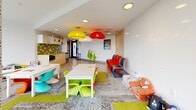 Located at 333 E 11th Ave in Vancouver's Mount Pleasant neighbourhood, this single-level children's amenity room is thoughtfully designed to provide a bright, engaging environment for young residents. The space is anchored by expansive floor-to-ceiling windows that flood the interior with natural light and offer sweeping elevated views of the surrounding Vancouver cityscape and neighbourhood rooftops.

The main play area features a patterned carpet with vibrant green and grey tones, complemented by a cheerful mix of colourful child-sized furniture, including bright lime green, teal, and orange chairs surrounding low activity tables. Eye-catching pendant lights in bold red, yellow, and white hang from the white recessed ceiling, adding a playful focal point to the room. Toy shelving units line the walls, keeping the space organized while remaining accessible for children.

A dedicated kitchenette runs along one wall, finished with light wood cabinetry, a chartreuse countertop, and a striking grey and lime green tile backsplash. It is equipped with a microwave and sink, providing a convenient refreshment area for supervising adults. A large whiteboard wall offers an additional creative outlet. The space also features a separate area with bean bags and soft plush toys near the windows, creating a cozy reading or relaxation nook with inspiring city views.