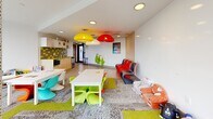 Located at 333 E 11th Ave in Vancouver's Mount Pleasant neighbourhood, this single-level children's amenity room is thoughtfully designed to provide a bright, engaging environment for young residents. The space is anchored by expansive floor-to-ceiling windows that flood the interior with natural light and offer sweeping elevated views of the surrounding Vancouver cityscape and neighbourhood rooftops.

The main play area features a patterned carpet with vibrant green and grey tones, complemented by a cheerful mix of colourful child-sized furniture, including bright lime green, teal, and orange chairs surrounding low activity tables. Eye-catching pendant lights in bold red, yellow, and white hang from the white recessed ceiling, adding a playful focal point to the room. Toy shelving units line the walls, keeping the space organized while remaining accessible for children.

A dedicated kitchenette runs along one wall, finished with light wood cabinetry, a chartreuse countertop, and a striking grey and lime green tile backsplash. It is equipped with a microwave and sink, providing a convenient refreshment area for supervising adults. A large whiteboard wall offers an additional creative outlet. The space also features a separate area with bean bags and soft plush toys near the windows, creating a cozy reading or relaxation nook with inspiring city views.