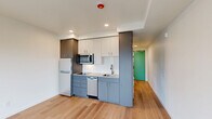Located at 1341 N Killingsworth St in Portland, Oregon, this efficiently designed studio offers a thoughtful layout that maximizes every square foot. The open-plan living area is anchored by light hardwood flooring that runs throughout, creating a warm and cohesive aesthetic. Large windows and a glass-paned balcony door flood the space with natural light while offering views of the surrounding Portland neighborhood, including a glimpse of a classic water tower. The balcony provides a convenient outdoor retreat.

The kitchen is outfitted with contemporary two-tone cabinetry in slate gray and white, a clean white countertop, stainless steel appliances including a microwave, dishwasher, and refrigerator, complemented by recessed lighting above. A dedicated nook houses a stacked in-unit washer and dryer, adding everyday convenience.

The bathroom features a sleek, modern design with a floating dark-cabineted vanity, an undermount sink, brushed nickel fixtures, a large mirror, and wood-look flooring that ties seamlessly to the rest of the unit. The overall ambiance is clean, bright, and move-in ready, reflecting contemporary urban residential design throughout.