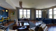This single-floor space makes a striking first impression with its thoughtfully designed living area, where abundant natural light pours in through a series of tall, large-pane windows dressed with patterned drapes. The elevated position of the space offers sweeping outdoor views of the surrounding neighborhood, adding a sense of openness and connection to the exterior environment.

Rich hardwood floors run throughout, grounding the space with warmth and texture. The living area is generously proportioned, arranged with a sophisticated mix of seating including a grey upholstered sofa, navy blue accent chairs, and a neutral sectional, all anchored by a dark wood coffee table atop a layered area rug. The color palette blends cool navy and grey tones with warm wood accents, creating a balanced and inviting ambiance.

A distinctive tiered chandelier with fringe detailing serves as a focal point overhead, adding an artful, eclectic touch to the space. Recessed ceiling lighting complements the natural light, ensuring the interior remains bright and welcoming. Decorative artwork on the walls adds personality and visual interest, completing the curated, contemporary aesthetic of this well-appointed space.