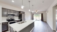 This well-appointed single-floor home offers a thoughtfully designed open-concept layout that seamlessly connects the kitchen and living areas. The kitchen serves as the centerpiece, featuring a large island with a striking white quartz countertop, an integrated sink, and pendant lighting overhead. Dark espresso-toned cabinetry provides rich contrast against the light countertops, while a full suite of stainless steel appliances — including a range, microwave, and refrigerator — adds a sleek, modern touch. The living area flows naturally from the kitchen and is bathed in natural light courtesy of expansive sliding glass doors that offer views of the surrounding greenery. A ceiling fan overhead enhances comfort, and a wall-mounted mini-split unit provides efficient climate control. Durable wood-look flooring runs throughout the main living spaces, adding warmth and continuity to the design. The bathroom is clean and functional, featuring a dark wood vanity with a white countertop, a well-lit mirror, and a tub-shower combination. A dedicated laundry room houses a full-size washer and dryer with overhead shelving for added storage. The space presents a cohesive, contemporary aesthetic that balances practicality with modern design sensibilities.