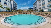 Located at 377 Santana Row in San Jose, California, this outdoor amenity space delivers a resort-caliber experience at the heart of one of Silicon Valley's most vibrant mixed-use destinations. The centerpiece is a generously sized oval swimming pool with crystal-clear blue water, elegantly framed by a decorative tile border and wide stone coping. The pool deck is extensively appointed with rows of cushioned chaise lounge chairs, providing ample space for sunbathing and relaxation.

A classically styled pergola with stone columns anchors the far end of the courtyard, serving as a shaded gathering point and adding an architectural focal point to the space. Tall, mature palm trees are strategically placed throughout the deck, enhancing the tropical ambiance and providing natural shade. A separate spa is situated along one side of the deck, complementing the main pool.

The courtyard is enclosed on multiple sides by the multi-story residential buildings, whose contemporary façades in grey and cream tones rise above the amenity level, offering a sense of privacy while allowing abundant natural light throughout the day. The overall design strikes a balance between Mediterranean-inspired architectural details and modern outdoor living, creating an inviting and well-equipped outdoor retreat suited to California's sunny climate.