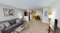 This single-floor residence offers an open-concept layout that seamlessly connects the living, dining, and kitchen areas into one cohesive, functional space. The living area is carpeted in a warm neutral tone and is furnished with a contemporary gray sofa, a glass-top coffee table, and a side table with a lamp, creating a comfortable and inviting atmosphere. Framed artwork on the walls adds a subtle decorative touch to the clean, light gray painted walls. A structural column divides the space visually without disrupting the open flow, leading the eye toward the dining and kitchen zones. The dining area accommodates a dark wood high-top table with matching bar-height chairs, ideal for casual meals. Just beyond, the kitchen features warm-toned wooden cabinetry, white appliances including a refrigerator and countertop coffee maker, and overhead flush-mount lighting that keeps the space bright and practical. A door visible near the column likely leads to additional rooms or storage, suggesting further private spaces within the unit. Throughout the entire space, the consistent carpeting, neutral color palette, and clean lines contribute to a cohesive, understated aesthetic that is both welcoming and easy to maintain.