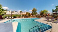 Located at 1820 Hickory Creek Ln in Columbus, Ohio, this outdoor amenity area centers around a generously sized swimming pool with crystal-clear blue water, accessible via stainless steel entry rails and steps. The pool deck is finished in smooth concrete and offers ample space for sunbathing, lined with multiple lounge chairs and seating areas positioned to take full advantage of the open sky and natural light.

The area features two distinct wooden pergola structures that provide shaded retreats for relaxation, complemented by additional covered gazebo seating with wicker furniture. A utility shed is conveniently situated near the pool perimeter, and a brick-accented community building with large windows borders one side of the space, adding architectural character to the setting.

The pool area is enclosed by a wrought iron fence, ensuring privacy and security, while well-maintained hedgerows, mature trees, and manicured landscaping create a lush, green backdrop throughout. Potted plants and shrubs further enhance the natural aesthetic. The surrounding residential buildings, featuring traditional gabled rooflines and chimneys, are visible beyond the tree line, reinforcing the community character of this outdoor space. Overall, the area offers a well-appointed, inviting environment designed for outdoor leisure and recreation.