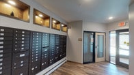 This single-floor common area functions as a mail and entry lobby, combining practical amenities with clean, contemporary design. The most prominent feature is a large bank of dark-finished individual mailboxes that spans a significant portion of one wall, providing organized mail access for multiple residents or tenants. The mailboxes are uniform in style, finished in a dark tone that contrasts with the light grey walls.

Above the mailbox bank, a series of warmly lit, wood-trimmed decorative wall niches add visual interest and a touch of character to an otherwise utilitarian space. These recessed displays are evenly spaced and illuminated from within, lending a refined aesthetic to the corridor.

The flooring throughout is a wood-look laminate in a warm, light tone, contributing to a clean and inviting atmosphere. A baseboard heating unit runs along the lower portion of the mailbox wall, indicating climate control provisions for year-round comfort.

At the far end of the hallway, a glass-paneled security door with a dark frame provides a controlled entry point, allowing natural light to filter into the space while maintaining building security. The overall ambiance is functional yet polished, reflecting a well-maintained and thoughtfully designed shared entryway.