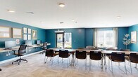 Located at 4795 Park Place Dr in Fort Wayne, Indiana, this single-floor community living room serves as a welcoming communal hub within a modern residential complex. The interior is defined by bold teal accent walls paired with crisp white ceilings and clean white baseboards, creating a vibrant yet polished atmosphere. Light-toned parquet-style carpeting runs throughout, contributing warmth and texture underfoot. Recessed lighting ensures consistent, even illumination across the space. Large windows dressed with sheer curtains invite an abundance of natural light, enhancing the open and airy feel of the room. The living area is thoughtfully arranged to accommodate both casual lounging and collaborative work, featuring a wall-mounted flat-screen television, comfortable seating, and a dining or meeting table with multiple chairs. A dedicated workstation along one wall provides additional utility. The space connects to a corridor leading to the building's interior. Outside, the complex presents a striking exterior finished in contrasting gray and navy blue horizontal siding over a brick base. A beautifully maintained central courtyard features manicured lawns, concrete pathways, garden benches, young trees, and well-tended landscaping beds — offering residents an inviting outdoor retreat directly accessible via French doors and ground-level entrances.