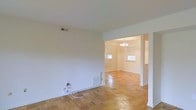This single-floor space showcases a clean, functional layout with notable architectural details that lend it a timeless character. The flooring throughout is a standout feature — classic parquet hardwood laid in a geometric pattern, adding warmth and visual interest underfoot. The neutral, white-toned walls keep the space feeling bright and airy, providing a versatile backdrop for any interior style.

The main living area flows seamlessly into an adjoining room via a wide, open doorway, creating a sense of connectivity and openness between spaces. The adjoining room benefits from natural light streaming in through multiple windows, enhancing the overall brightness of the interior. A chandelier with a traditional multi-arm design serves as an elegant focal point in this connected space, adding a refined touch to the otherwise understated aesthetic.

Practical details include ceiling-mounted HVAC vents, a floor-level heating register, and wall outlets positioned conveniently throughout. The consistent flooring and wall finishes across both visible areas contribute to a cohesive, unified feel. Overall, the space presents a bright, well-connected interior with classic design elements and a welcoming ambiance.