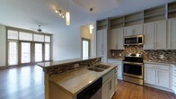 This single-floor residence showcases a thoughtfully designed open-concept layout that seamlessly connects the kitchen and living areas. The kitchen serves as the centerpiece of the space, featuring a large central island with granite countertops and an integrated sink, ideal for both meal preparation and casual dining. Soft gray shaker-style cabinetry lines the walls, complemented by open upper shelving that adds a modern, airy feel. A mosaic tile backsplash in warm earth tones adds texture and visual interest behind the stainless steel range and built-in microwave. Pendant lighting and a track light fixture illuminate the kitchen, enhancing its contemporary aesthetic. The generous counter space and full-height cabinetry provide ample storage throughout. Rich, dark hardwood flooring flows continuously through the kitchen and into the adjoining living area, grounding the space with warmth. Large windows allow natural light to filter in, brightening the open living zone. A recessed ceiling detail above the kitchen adds architectural depth, while a neatly integrated entryway and interior doors contribute to the overall cohesive design. This space balances functional design with stylish, modern finishes throughout.