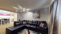 This single-floor space presents a cohesive and contemporary design aesthetic throughout. The living area serves as the focal point, anchored by a generously sized black leather sectional sofa that offers ample seating and a sleek, modern feel. Accent pillows in red, black, and white geometric patterns add a vibrant contrast to the dark upholstery, lending a pop of personality to the space.

The walls are finished in a cool neutral grey tone, creating a calm and sophisticated backdrop that complements the bold furnishings. A striking abstract canvas artwork in grey, white, and metallic tones adorns the wall, reinforcing the modern aesthetic. Decorative star-burst wall accents contribute an additional layer of visual interest.

Lighting plays a key role in the ambiance of this space. A multi-arm arc floor lamp provides functional task lighting while also serving as a design element, and recessed ceiling lighting casts a warm, even glow. Decorative light reflections on the ceiling add a unique and dynamic quality to the room's atmosphere.

A glass-topped coffee table anchors the seating area, while sculptural red decorative pieces on its surface echo the accent colors present throughout. A corridor visible in the background suggests additional rooms beyond, completing this well-appointed and thoughtfully styled single-level space.