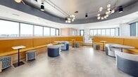 Located in Minneapolis's University Avenue corridor, this thoughtfully designed amenity space occupies a generous upper floor, offering a sophisticated blend of industrial and contemporary aesthetics. Polished concrete floors extend throughout the open-plan interior, complemented by exposed concrete columns and visible ductwork that lend the space an urban, loft-like character.

The interior is organized into distinct seating zones, each with its own personality. A standout curved banquette upholstered in warm golden-yellow wood wraps around a central gathering area beneath a dramatic arched ceiling detail, creating an inviting social hub. Nearby, teal and natural rattan lounge chairs, modular soft seating, and bar-height counters provide flexible arrangements for both casual interaction and focused use. Decorative globe pendant lighting and bold pendant fixtures add warmth and visual interest throughout.

Floor-to-ceiling windows and large black-framed glazing flood the interior with natural light while framing expansive views of the Minneapolis skyline and surrounding neighborhoods. An adjacent rooftop terrace, clad in clean large-format pavers and dark brick, extends the experience outdoors with ample lounge seating, fire pit tables, and unobstructed panoramic city views — ideal for gathering under open skies.