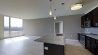 This single-floor space is designed with a contemporary open-concept layout that seamlessly integrates the kitchen and living areas. The kitchen is anchored by a substantial island finished in granite, featuring subtle grey and white veining that adds visual interest and texture. The dark-toned cabinetry of the island provides a striking contrast against the light-colored flooring, which extends throughout the main living area. A stainless steel sink is embedded in the island, adding functional elegance to the space. Overhead, two cylindrical pendant lights hang at varying heights, contributing a modern design element while providing focused task lighting. The ceilings are clean and well-proportioned, accommodating recessed lighting fixtures in addition to the pendants. The walls are finished in a neutral grey tone, lending a contemporary and cohesive aesthetic. A doorway visible in the background leads to an adjacent room, suggesting a well-connected floor plan. Large floor-to-ceiling windows along one wall allow generous amounts of natural light to fill the space, creating a bright and airy atmosphere. The overall ambiance is modern, clean, and functional.