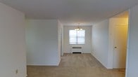 This single-floor residential space offers a clean, understated layout designed for comfortable everyday living. The interior is characterized by bright white walls and neutral-toned wall-to-wall carpeting that runs throughout, creating a cohesive and welcoming atmosphere. The open living and dining area is a standout feature of the space, bathed in natural light that streams through a large window framed by the exterior greenery beyond. A classic chandelier hangs above the dining area, adding a traditional decorative touch to the otherwise minimalist design. A radiator beneath the window provides efficient heating, a nod to timeless residential infrastructure. The open layout between the living and dining areas allows for flexible furniture arrangements and promotes a sense of spaciousness. A partial wall divides the main living space from an adjoining area, contributing to a defined yet open floor plan. The simple, well-maintained finishes throughout — from the smooth white ceilings to the clean baseboards — reflect a neutral palette that serves as an ideal canvas for personalization. Overall, the space presents a functional and light-filled environment with classic residential character.