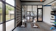 Located at 6925 Roswell Rd in Sandy Springs, GA, this single-floor fitness center is thoughtfully designed to support a wide range of workout routines. The facility is divided into distinct zones, each tailored to a specific training style.

The spin studio is a standout feature, enclosed by sleek floor-to-ceiling steel-framed glass partitions that provide both visual openness and acoustic separation. The room is outfitted with multiple stationary cycling bikes and benefits from generous natural light streaming through large windows that offer views of the surrounding greenery and an adjacent outdoor balcony area.

Adjacent to the spin studio is the main weight room, visible through the glass walls and featuring a comprehensive selection of strength training machines and free weights, all set on hardwood-style flooring beneath recessed lighting.

A dedicated functional training room rounds out the facility, equipped with an extensive multi-tiered equipment rack stocked with stability balls, medicine balls, kettlebells, resistance tools, and BOSU trainers. A wall-mounted exercise mat and a large flat-screen display — integrated with a fitness app platform — make this space ideal for guided, self-directed workouts. The consistent use of neutral tones, carpet flooring, and modern finishes throughout creates a clean, professional atmosphere conducive to focused training.