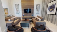 This elegantly designed single-floor amenity space seamlessly blends comfort and sophistication across several distinct areas. The centerpiece lounge features soaring ceilings and a dramatic floor-to-ceiling horizontal wood-slat accent wall housing a large flat-screen television. Plush grey sofas, curved armchairs with warm wood frames, and a tufted ottoman are arranged atop a patterned area rug, creating an inviting gathering space. Curated artwork and decorative blue glass accents lend a polished, contemporary character throughout.

The kitchen area is a standout, anchored by a sleek white waterfall island with bar seating for several guests. Dark walnut cabinetry frames a striking geometric-patterned tile backsplash, while stainless steel appliances, including a built-in wall oven and French door refrigerator, provide full functionality. An eye-catching sculptural branch chandelier cascades overhead, adding artistic flair.

A private meeting or dining room features a substantial solid wood table, upholstered banquette seating, dark bentwood chairs, herringbone wood-look flooring, and an industrial-style multi-arm chandelier, offering an intimate setting for small gatherings. Additional areas visible within the space include a game room with a billiard table. Large-format porcelain tile flooring and textured wallcoverings with a vertical stripe motif unify the space with a refined, modern aesthetic throughout.