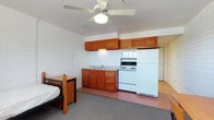 This single-floor studio-style unit offers an open-plan layout that efficiently combines living, sleeping, and kitchen functions within one shared space. The kitchenette lines one wall and is equipped with warm-toned wooden cabinetry — both upper and lower — alongside a white electric range, over-range exhaust hood, full-size refrigerator, and a single-basin sink. The cabinetry provides ample storage while lending a touch of warmth to the otherwise utilitarian setting. The walls are constructed of painted white brick, giving the space a solid, institutional character that is clean and straightforward. Flooring transitions from a neutral carpet in the living and sleeping area to a hard surface near the kitchen zone, offering practical differentiation between spaces. A ceiling fan with an integrated light fixture is centrally mounted, providing both air circulation and ambient lighting. A desk and bed frame are visible, confirming the dual-purpose nature of the layout. A doorway visible in the background suggests access to an additional room, potentially a bathroom. The overall ambiance is functional and no-frills, suited for straightforward, efficient living.
