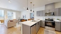 This thoughtfully designed 2-bedroom, 2-bathroom single-floor residence offers a cohesive modern aesthetic throughout. The open-concept kitchen and living area serves as the heart of the home, featuring gray shaker-style cabinetry with matte black hardware, a white quartz peninsula with bar seating, stainless steel appliances including a full-size refrigerator, range, and microwave, and stylish pendant lighting overhead. Light wood-tone flooring unifies the space and extends into the living area, which benefits from large windows and a glass door that invite generous natural light and provide access to an outdoor balcony. The living room offers a bright, comfortable atmosphere with clean white walls and views to the exterior. Both bedrooms are carpeted and well-lit by windows with outdoor views, providing a calm and restful environment. The primary bedroom includes a dedicated walk-in closet outfitted with wire shelving and hanging rods for organized storage. The bathrooms are finished with gray vanity cabinetry, white countertops, and wood-tone flooring, with one bath featuring a full tub-shower combination. Consistent use of neutral tones, modern fixtures, and quality finishes throughout creates a clean, contemporary living environment.