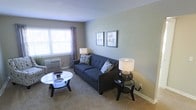 This single-floor space features a thoughtfully arranged living room that serves as its central gathering area. The room is finished with wall-to-wall carpeting in a warm neutral tone, complemented by soft sage-green walls that create a calm, understated ambiance. A window along one wall allows natural light to filter in, dressed with decorative curtains that add a touch of softness to the space. A window-mounted air conditioning unit provides climate control.

The living room is furnished with a tufted navy blue sofa accented by patterned throw pillows, positioned to face a round glass-topped coffee table with a dark frame. Two side lamps with globe-style bases and white shades provide warm ambient lighting. A pair of framed prints mounted on the wall add visual interest and a modest decorative touch. The clean white baseboards contrast neatly against the carpeted floor, contributing to a tidy, well-maintained appearance. Overall, the space offers a compact yet comfortable layout suited for everyday living.