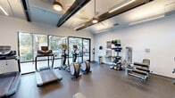 This fitness center at Arrive Rolling Oaks is a thoughtfully designed, single-level workout facility that combines functionality with a contemporary aesthetic. The space is divided into distinct workout zones, allowing for a comprehensive range of exercise options. Large floor-to-ceiling windows and glass doors along one wall flood the interior with natural light while offering views of the surrounding outdoor greenery, creating an open and inviting atmosphere. A glass-door entry provides direct access to the exterior courtyard area.

The cardio zone is well-stocked with treadmills, elliptical trainers, and upright bikes, all positioned to take advantage of the natural light from the expansive window wall. Overhead, exposed black structural beams add an industrial contrast to the clean white walls, while ceiling fans and linear LED lighting ensure a comfortable environment during workouts.

The strength training area features a multi-station cable machine, a flat workout bench, and a full rack of dumbbells arranged in front of a large wall mirror. Upper clerestory windows further brighten this section. A dedicated accent wall painted in bold blue anchors a separate strength zone equipped with a treadmill and wall-mounted flat-screen television. Rubber flooring throughout provides durability and safety. A hydration station and sanitizer dispenser complete the practical amenities.