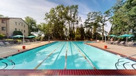Located at 101 Clifton Park Cir in Wilmington, Delaware, this outdoor aquatic facility centers around a generously sized lap pool with clearly marked swim lanes, making it well-suited for both recreational swimming and structured exercise. The pool features classic brick-red coping along its edges and is surrounded by a broad concrete deck that provides ample space for movement and socializing.

The surrounding deck is furnished with lounge chairs and shaded umbrella seating areas, offering comfortable spots for relaxation. A dedicated patio area with a round table and chairs provides additional outdoor gathering space, enclosed by a stone retaining wall and wrought iron fencing that lend a sense of structure and security to the environment.

Several small support buildings line the perimeter of the pool area, appearing to house facilities such as changing rooms, storage, and maintenance spaces. Their neutral siding with green trim gives the complex a cohesive, well-maintained appearance.

The entire facility is embraced by a canopy of mature trees that provide natural shade and a lush, green backdrop, contributing to a tranquil and inviting outdoor atmosphere. The combination of open sky, verdant surroundings, and well-organized amenities makes this a functional and appealing community recreational destination.