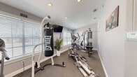 Located at The Vista in Winter Park, Florida, this dedicated fitness center offers a well-equipped workout space designed to support a comprehensive exercise routine. The room features neutral gray walls, durable carpet flooring, a textured ceiling with recessed lighting, and multiple windows dressed with white horizontal blinds that allow ample natural light to fill the space.

The fitness center is outfitted with a variety of equipment to accommodate diverse training needs. Cardio options include a treadmill and an elliptical machine, while a multi-station cable weight machine provides a wide range of strength-training exercises. An Everlast heavy punching bag on a freestanding stand adds a boxing and functional training element to the room. A wall-mounted flat-screen television is positioned for viewing during workouts.

Informational posters, workout charts, and fitness center rules are displayed on the walls, keeping users informed and oriented. A wall clock and motivational signage contribute to the purposeful atmosphere of the space. Entry to the fitness center is through a glass-paned door that opens to the surrounding outdoor community grounds, reinforcing the connection to the broader property. The overall ambiance is clean, bright, and functional, making it a practical amenity for residents of The Vista community.