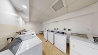 This communal laundry facility is a practical, efficiently organized space designed to accommodate multiple users simultaneously. The room houses a substantial number of top-loading and front-loading washing machines along with several dryers, all finished in white and arranged along the walls to maximize usable floor space. Each machine is paired with coin-operated or card-reader payment panels mounted on the wall, offering straightforward operation.

The flooring throughout is classic terrazzo, featuring a warm speckled tan tone that is both durable and easy to maintain. A drop ceiling with recessed lighting and integrated ventilation panels provides even, functional illumination across the room. The walls are painted a clean, bright white, contributing to the overall sense of cleanliness and order.

A small round folding table is positioned near the perimeter of the room, offering a convenient surface for sorting or folding laundry. A radiator unit is also present, providing heat to the space. A utility sink along one wall adds further functionality.

Access to the room is through a glass-paneled door marked with a "Please Keep Door Closed" notice, opening onto a corridor finished with matching terrazzo flooring and white tile walls. A fire extinguisher is mounted in the hallway, reflecting attention to safety standards throughout the facility.
