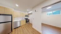Located at 3417 Harbor Ave SW in Seattle's West Seattle neighborhood, this efficiently designed unit offers a clean, modern aesthetic throughout its single-floor layout. Warm hardwood-style flooring runs continuously across all areas, creating a cohesive and inviting atmosphere.

The kitchen is a standout feature, equipped with a full suite of stainless steel appliances including a refrigerator, electric range, over-the-range microwave, and dishwasher. Light natural wood cabinetry extends from floor to ceiling, providing generous storage, while white countertops and a stainless steel sink complete the crisp, functional design. Recessed lighting overhead keeps the space well-lit and open.

The full bathroom is finished in a clean, minimalist style, featuring a white vanity with under-sink storage, a toilet, and a walk-in shower stall with chrome fixtures and a built-in shelf. The light-toned flooring carries through here as well, maintaining visual consistency.

Two closets offer practical storage solutions, one of which features wire shelving systems that maximize organization. A connecting hallway links the spaces efficiently, with a recessed utility panel integrated neatly into the wall. A window admits natural light into the living area, brightening the overall space. The unit's design prioritizes clean lines, functional storage, and a modern, neutral palette throughout.