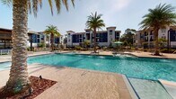 Located at 8890 Ransley Station Blvd in Pensacola, Florida, this outdoor amenity space delivers a resort-style experience set against the backdrop of a modern multi-story residential community finished in bold navy blue and crisp white. At the heart of the space is a generously sized swimming pool with built-in water features and in-pool lounge platforms, designed for both relaxation and recreation. The pool deck is finished in warm, textured concrete and is dotted with tall palm trees that evoke a true Florida coastal atmosphere under abundant natural sunlight. Surrounding the pool, ample seating areas are outfitted with lounge chairs and umbrella-shaded tables, offering residents a variety of options for outdoor leisure. A substantial wood-framed pergola on one side of the deck provides a shaded gathering area complete with additional seating and an outdoor kitchen or grill station. Manicured landscaping with lush greenery, ornamental shrubs, and mulched garden beds lines the perimeter walkways and black aluminum fencing, creating a polished and well-maintained environment. Hammocks are thoughtfully placed throughout the grounds for added relaxation. Wide, smooth pathways connect all areas of the amenity space, making it accessible and easy to navigate throughout the community.