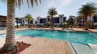Located at 8890 Ransley Station Blvd in Pensacola, Florida, this outdoor amenity space delivers a resort-style experience set against the backdrop of a modern multi-story residential community finished in bold navy blue and crisp white. At the heart of the space is a generously sized swimming pool with built-in water features and in-pool lounge platforms, designed for both relaxation and recreation. The pool deck is finished in warm, textured concrete and is dotted with tall palm trees that evoke a true Florida coastal atmosphere under abundant natural sunlight. Surrounding the pool, ample seating areas are outfitted with lounge chairs and umbrella-shaded tables, offering residents a variety of options for outdoor leisure. A substantial wood-framed pergola on one side of the deck provides a shaded gathering area complete with additional seating and an outdoor kitchen or grill station. Manicured landscaping with lush greenery, ornamental shrubs, and mulched garden beds lines the perimeter walkways and black aluminum fencing, creating a polished and well-maintained environment. Hammocks are thoughtfully placed throughout the grounds for added relaxation. Wide, smooth pathways connect all areas of the amenity space, making it accessible and easy to navigate throughout the community.