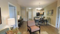 This single-floor residence features an open-concept layout that seamlessly connects the living, dining, and kitchen areas into one cohesive, functional space. Hardwood-style flooring flows continuously throughout, lending warmth and visual continuity across the entire unit.

The living area is furnished with comfortable seating, including a patterned accent chair and a floral sofa, set atop a textured area rug. A console table with a lamp anchors one side of the space, adding a decorative element near what appears to be a mirrored closet or entry nook.

The dining area sits centrally within the open floor plan, featuring a rectangular table with dark chairs accommodating multiple diners. Overhead, pendant lights on a central fixture provide focused illumination, enhancing the sense of definition within the shared space.

The kitchen is positioned at the rear and is equipped with light wood cabinetry, a black refrigerator, and countertop workspace. Additional pendant lighting above the kitchen area reinforces a coordinated lighting scheme throughout.

White interior doors and neutral beige walls contribute to a clean, bright, and welcoming atmosphere. The layout offers an efficient use of space with clear sightlines from the living area through to the kitchen.