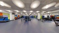 Located at 800 S Wells St in Chicago's South Loop, this expansive fitness center — branded as River — offers a comprehensive, single-floor workout facility thoughtfully designed to accommodate a wide range of training styles. The space is characterized by a bold, energizing color palette of cobalt blue, lime green, orange, and purple, applied across accent walls and structural columns to create distinct zones throughout the facility. Durable rubber flooring in blue and gray tones covers the main workout areas, while synthetic turf defines the functional training zone.

The cardio area features spin bikes, treadmills, stair climbers, and rowing machines arranged across open floor space, complemented by a curved indoor track with turf sections for agility and sled work. A dedicated strength training zone houses power racks, cable machines, free weights, and benches, set against deep blue walls. A separate room with glass partitions accommodates cycling classes and additional cardio equipment. Unique oval pendant light fixtures and exposed industrial ceiling infrastructure add architectural character throughout. A water station and motivational wall art — including a "Choose Happy" sign — contribute to the welcoming atmosphere. Glass walls connect certain zones, maintaining an open, airy feel across the full facility.