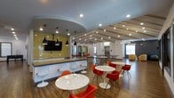 This single-story clubhouse offers a dynamic and well-appointed communal space designed to foster social interaction and relaxation. The open-concept layout seamlessly connects multiple activity zones, creating an inviting atmosphere throughout.

The main lounge and dining area is anchored by sleek marble-top round tables paired with bold red molded chairs, complemented by white tulip-style seating along a built-in counter. Striking black pendant lights hang overhead, while recessed lighting illuminates the vaulted, beam-detailed ceiling that adds architectural character to the space. The flooring features warm wood-tone planks that enhance the modern aesthetic.

A kitchenette area is finished with eye-catching yellow-green subway tile backsplash and is equipped with a mounted flat-screen TV, making it ideal for casual gatherings. Beyond the main seating area, additional recreational space is visible, featuring game tables and comfortable lounge seating near large windows that invite natural light. The overall ambiance balances energetic design elements with functional versatility, making this clubhouse well-suited for a variety of community activities.