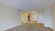 This single-floor unit offers a straightforward, functional layout designed to maximize usable living space. The main living area is generously proportioned, finished with wall-to-wall beige carpeting that extends throughout, lending a cohesive and comfortable feel underfoot. Walls are painted in a soft, neutral tone, contributing to a clean and bright overall ambiance. Overhead, flush-mount ceiling lighting provides consistent illumination, while HVAC vents are positioned throughout the ceiling to ensure efficient climate control.

The kitchen is open and directly accessible from the main living area, separated by a structural column that subtly defines each space without fully enclosing them. It features warm-toned wooden cabinetry, dark countertops, and stainless steel appliances, including a range, microwave, and refrigerator, offering a practical cooking environment. The kitchen floor transitions to a lighter hard surface, clearly distinguishing it from the carpeted living space.

From the living area, doorways lead to additional rooms, including what appears to be a hallway accessing further living quarters. A louvered closet door is visible near one of the hallway entrances, indicating dedicated storage. The entry door, finished in white, is positioned along one wall, providing a clear and accessible point of entry into the unit. Overall, the space presents a clean, move-in-ready environment with a neutral palette suitable for a variety of interior styles.