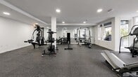 Located at 5777 177B St in Surrey, BC, this dedicated fitness facility offers a thoughtfully designed exercise space spread across a single floor. The room is finished with a clean, modern aesthetic — crisp white walls and structural columns contrast with dark speckled rubber flooring that spans the entire area, providing both durability and slip resistance underfoot.

The strength training zone is generously outfitted with a multi-station cable machine, squat racks, a dumbbell rack with a full range of free weights, barbells, and adjustable benches, giving users everything needed for a comprehensive weight training session. A floor-to-ceiling mirror along one wall enhances the sense of depth and allows for form checking during workouts.

The cardio section benefits from exceptional natural light, with large floor-to-ceiling windows overlooking a landscaped outdoor courtyard and surrounding residential buildings — creating an open, energizing atmosphere. Treadmills, a stair climber, and an elliptical trainer are positioned to take full advantage of these views.

A built-in white cubby storage unit provides convenient personal item storage, while recessed ceiling lighting ensures the space remains bright and well-lit throughout. An accessible washroom is also conveniently located nearby. Overall, the facility is clean, functional, and well-maintained, offering a complete fitness experience in a bright and modern environment.