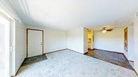 This single-floor space presents an open and functional layout designed for comfortable everyday living. The main living area is finished with light-colored wall-to-wall carpeting that lends a warm, soft feel underfoot, while dark tile flooring defines a transitional zone near the entryway, creating a natural boundary between spaces. The front entry door features classic wood trim detailing that adds a subtle traditional touch to the otherwise neutral interior.

The open floor plan flows seamlessly into an adjoining area, distinguished by the same dark tile flooring and equipped with a ceiling fan fitted with a light fixture, providing both ventilation and ambient lighting. This connected space offers views into a further room that appears to feature built-in shelving or cabinetry, suggesting additional storage or organizational potential.

Throughout the space, white walls and a textured ceiling contribute to a clean, understated aesthetic. Baseboard trim runs consistently along the walls, adding a finished quality to the interior. The overall ambiance is bright, open, and neutral, offering a versatile canvas well-suited for a variety of furnishing styles and functional arrangements.