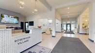 Located in Powell, Ohio, the Rosewood Village clubhouse is a thoughtfully designed community facility that offers a refined and welcoming atmosphere across a single, generously sized floor. The grand entrance lobby immediately impresses with soaring vaulted ceilings, elegant crystal chandeliers, and large-format light tile flooring. A stylish reception desk bearing the Rosewood Village branding greets visitors, while an adjacent lounge area with a round dining table and modern pendant lighting provides an inviting gathering space.

The business center features a long workstation lined with desktop computers set against a striking botanical-patterned accent wall with globe pendant lights, offering a functional yet visually appealing workspace. Nearby, a fully equipped kitchen with stainless steel appliances, white cabinetry, and bar-height seating beneath globe pendant lights serves as a convenient amenity for residents.

The media room is a standout feature, offering a wall-mounted flat-screen TV, plush curved seating, and soft carpeting for a comfortable viewing experience. A separate lounge area with a floor-to-ceiling stacked stone fireplace, multiple seating arrangements, an additional mounted TV, and abundant natural light from large windows creates a warm and sophisticated retreat throughout the space.