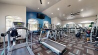 Located at 5151 E Guadalupe Rd in Phoenix, AZ, this fitness center offers a well-appointed, single-floor exercise space designed to support a comprehensive workout experience. The room features soaring vaulted ceilings that create an open, airy atmosphere, complemented by an abundance of natural light streaming through multiple large arched windows that frame views of the surrounding outdoor landscape.

The interior design is defined by a bold blue accent wall that adds energy and visual interest to the space, contrasting with the crisp white walls and ceilings. Recessed lighting and a ceiling fan ensure the room remains well-lit and comfortable during use. Patterned, multi-tone carpet tiles cover the floor throughout, adding texture and a modern aesthetic.

The fitness center is comprehensively equipped with a wide variety of cardio machines, including treadmills, ellipticals, and stationary bikes, arranged to maximize floor space and ease of movement. A dedicated strength training area features cable machines, a squat/power rack with free weights, medicine balls, and additional resistance equipment. Full-length mirrors along one wall enhance the sense of space and allow users to monitor their form. A wall-mounted flat-screen television provides entertainment during workouts, rounding out this fully functional and thoughtfully designed fitness facility.