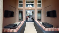 This single-floor outdoor terrace lounge offers a well-appointed gathering space that seamlessly blends indoor comfort with open-air living. The space is anchored by modular wicker sectional seating dressed in warm earth-toned striped cushions and decorative pillows, arranged to encourage relaxed conversation and entertainment. A wall-mounted flat-screen television is integrated directly into the textured stucco wall, supported by neatly routed cabling, making it ideal for outdoor viewing. Dramatic floor-to-ceiling red drapery panels add a bold, lounge-like aesthetic, framing the transition between the sheltered and open portions of the terrace.

The open-air section features a glass-enclosed fire pit as a central focal point, flanked by additional wicker seating and shaded by a large terracotta market umbrella. Tall columnar architectural details lend a Mediterranean-inspired character to the structure, while views of surrounding palm trees and the urban streetscape provide a lively backdrop. The entry façade features large glass double doors beneath expansive multi-pane windows, flooding the interior threshold with natural light. The overall ambiance is sophisticated yet approachable, suited for both casual lounging and social gatherings.