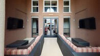 This single-floor outdoor terrace lounge offers a well-appointed gathering space that seamlessly blends indoor comfort with open-air living. The space is anchored by modular wicker sectional seating dressed in warm earth-toned striped cushions and decorative pillows, arranged to encourage relaxed conversation and entertainment. A wall-mounted flat-screen television is integrated directly into the textured stucco wall, supported by neatly routed cabling, making it ideal for outdoor viewing. Dramatic floor-to-ceiling red drapery panels add a bold, lounge-like aesthetic, framing the transition between the sheltered and open portions of the terrace.

The open-air section features a glass-enclosed fire pit as a central focal point, flanked by additional wicker seating and shaded by a large terracotta market umbrella. Tall columnar architectural details lend a Mediterranean-inspired character to the structure, while views of surrounding palm trees and the urban streetscape provide a lively backdrop. The entry façade features large glass double doors beneath expansive multi-pane windows, flooding the interior threshold with natural light. The overall ambiance is sophisticated yet approachable, suited for both casual lounging and social gatherings.