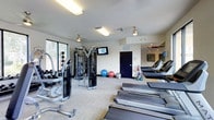 This fitness center offers a thoughtfully arranged workout space designed to accommodate a variety of exercise routines. The room features neutral-toned walls and a light carpet floor, contributing to a clean and functional atmosphere. Overhead fluorescent and pendant lighting ensures the space is well-illuminated throughout.

On the cardio side, multiple treadmills are lined up along one side of the room, providing ample capacity for users. A multi-station cable machine serves as a centerpiece for strength training, complemented by a selection of free weights and additional resistance equipment positioned nearby. Exercise balls in various sizes and a rack of kettlebells add versatility for functional fitness training.

A wall-mounted flat-screen television is positioned centrally, offering entertainment during workouts. A large window along one wall brings in natural light and provides views of surrounding greenery, enhancing the overall ambiance. A navy blue door leads to an adjacent area, and decorative wall art adds a finishing touch. The space is efficiently organized to maximize usability while maintaining an open, accessible feel.