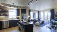 This single-floor space showcases a seamlessly integrated open-concept layout that connects the kitchen and living area into one cohesive, functional environment. The kitchen is outfitted with dark espresso cabinetry that extends to the ceiling, providing ample storage while contributing to the space's sleek aesthetic. A stainless steel French-door refrigerator anchors the kitchen, complemented by a undermount sink positioned beneath a bright window. A dark-finished breakfast bar with bar seating serves as a natural divider between the kitchen and living area, offering a casual dining or entertaining option.

Track lighting runs throughout the ceiling, providing versatile, adjustable illumination across both the kitchen and living zones. Light hardwood flooring adds warmth and continuity throughout the space. The living area is generously proportioned, featuring a sectional sofa, a glass-top coffee table, and a large area rug anchoring the seating arrangement. Multiple large windows and a glass patio door flood the interior with natural light and offer views of the surrounding landscape. Sheer and drape window treatments frame each opening, adding texture and depth. The overall ambiance is modern, sophisticated, and inviting.