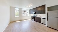 Located at 1943 Dorchester Ave in Boston's Dorchester neighborhood, this single-floor unit offers a thoughtfully designed open-plan layout that maximizes usable space. The main living area features light-toned hardwood-style flooring and crisp white walls, creating a bright and airy atmosphere. A wall-mounted mini-split air conditioning unit provides year-round climate control. Large windows along the exterior wall invite generous amounts of natural light and offer views of the surrounding neighborhood streetscape.

The kitchen is seamlessly integrated into the living space and is outfitted with rich, dark wood cabinetry extending from floor to ceiling, offering ample storage. Stainless steel appliances — including a range, over-the-range microwave, dishwasher, and refrigerator — complement the clean white countertops, giving the kitchen a sleek, modern aesthetic. The light flooring continues throughout, unifying the open space.

The bathroom is finished in a clean, contemporary style with white walls, wood-look flooring, and a full tub/shower combination equipped with an accessibility grab bar. A dark wood vanity with a white countertop and an integrated sink rounds out the well-appointed space. A closet area provides additional storage. The overall design is modern, functional, and move-in ready.
