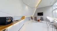 This single-floor space makes an immediate impression with its bold, futuristic architectural design. The interior is defined by dramatically angled white wall and ceiling panels that create a dynamic, geometric aesthetic throughout. Seamlessly integrated LED strip lighting traces the angular lines of the ceiling, casting a warm, ambient glow that enhances the sense of depth and dimension.

The kitchenette area is thoughtfully designed, featuring clean white cabinetry paired with a rich warm-toned wood countertop that provides a striking natural contrast to the otherwise all-white environment. A stainless steel sink and modern faucet complete the functional setup, while under-cabinet lighting adds both practicality and visual warmth.

Adjacent to the kitchenette, a contemporary lounge area offers a comfortable gathering space. A selection of modern swivel chairs in neutral grey tones, complemented by an accent chair with a warm patterned upholstery, are arranged to encourage conversation and collaboration. A wall-mounted flat-screen display serves as the focal point of this seating zone, making it well-suited for presentations or media viewing.

The seamless white flooring unifies the space and amplifies the bright, clean atmosphere created by the architectural lighting. Overall, the space conveys a highly curated, design-forward environment ideal for professional or creative use.
