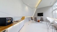 This single-floor space makes an immediate impression with its bold, futuristic architectural design. The interior is defined by dramatically angled white wall and ceiling panels that create a dynamic, geometric aesthetic throughout. Seamlessly integrated LED strip lighting traces the angular lines of the ceiling, casting a warm, ambient glow that enhances the sense of depth and dimension.

The kitchenette area is thoughtfully designed, featuring clean white cabinetry paired with a rich warm-toned wood countertop that provides a striking natural contrast to the otherwise all-white environment. A stainless steel sink and modern faucet complete the functional setup, while under-cabinet lighting adds both practicality and visual warmth.

Adjacent to the kitchenette, a contemporary lounge area offers a comfortable gathering space. A selection of modern swivel chairs in neutral grey tones, complemented by an accent chair with a warm patterned upholstery, are arranged to encourage conversation and collaboration. A wall-mounted flat-screen display serves as the focal point of this seating zone, making it well-suited for presentations or media viewing.

The seamless white flooring unifies the space and amplifies the bright, clean atmosphere created by the architectural lighting. Overall, the space conveys a highly curated, design-forward environment ideal for professional or creative use.