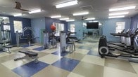 This fitness center offers a functional and organized space for residents or members to maintain an active lifestyle. The room is finished with a bold blue and cream checkered floor, adding a distinct visual character to the space. Walls are painted in a calming blue tone, contributing to an energized yet focused atmosphere.

The facility is equipped with a selection of strength-training machines, including cable and resistance units, as well as a rowing machine and an elliptical trainer, providing options for both upper and lower body workouts. A stability ball and additional free weights, visible in the mirrored wall reflection, further expand the available equipment.

A large flat-screen television is mounted on the wall, offering entertainment during workouts. The mirrored wall not only makes the space feel larger but also allows users to monitor their form. Overhead fluorescent lighting ensures the room is well-lit throughout, and a ceiling fan aids in air circulation. A window along one wall introduces natural light, enhancing the overall comfort of the environment.