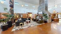 Located at 211 North Ave in Athens, GA, The Lodge is a generously sized single-floor gathering space that masterfully blends rustic warmth with mid-century modern design. The centerpiece of the main hall is a soaring vaulted ceiling that rises dramatically overhead, drawing the eye upward toward a striking antler chandelier and rows of clerestory windows that flood the space with natural light. A monumental stacked-stone fireplace anchors one end of the great room, surrounded by plush leather seating, patterned area rugs, and an assortment of lounge furniture arranged for both intimate conversation and open socializing. Warm terracotta-toned polished concrete floors run throughout the main areas, complemented by rich wood-paneled walls that lend an inviting, lodge-like character. A decorative hand-carved totem pole adds a bold artistic focal point to the expansive open floor plan. Adjacent to the main hall, a more intimate lounge area offers a contrast in atmosphere — featuring slate-blue vertical shiplap walls, a rich wood-planked ceiling, carpeted floors, and a large flat-screen display, ideal for private screenings or relaxed gatherings. A hallway connects the various living areas, which also include a kitchenette and additional flexible spaces. Lush indoor plants are thoughtfully placed throughout, enhancing the organic, nature-inspired ambiance of this distinctive communal retreat.
