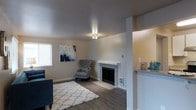 This single-story space presents a well-appointed living area designed with both comfort and functionality in mind. The living room serves as the focal point, anchored by a classic fireplace with a clean white surround and tile detailing, providing a warm gathering spot. The room features durable hardwood-style flooring in a rich, medium-toned finish that runs throughout, adding warmth and visual continuity to the space.

Neutral, light-toned walls create a bright and airy feel, complemented by a textured ceiling with a central flush-mount light fixture that evenly illuminates the room. A window fitted with horizontal blinds allows for natural light and privacy control. The open layout flows naturally into an adjacent area, partially visible through a columned partition, hinting at a connected kitchen or dining space with countertop details.

The overall ambiance is clean, modern, and move-in ready, with a understated color palette that provides a versatile backdrop for a variety of interior styles. The space is practical and well-structured, making it suitable for comfortable everyday living.