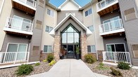 This multi-story residential building presents a well-designed exterior that blends contemporary and traditional architectural elements. The standout feature of the building is its grand entrance, framed by natural stone pillars and topped with a dramatic peaked glass facade that draws the eye upward and allows ample natural light into the entryway. A wide concrete walkway leads visitors directly to the double glass doors, which are flanked by decorative planters and neatly arranged landscaping, adding a welcoming and polished curb appeal.

The building's exterior is clad in neutral-toned horizontal siding, accented by white trim that lends a clean, classic look. Multiple private balconies extend from the upper floors, providing residents with outdoor space and views of the surrounding area. The balconies feature white railings, complementing the overall color palette of the structure.

The landscaping surrounding the entrance incorporates a mix of ornamental grasses, shrubs, and seasonal plantings set within a low-maintenance rock ground cover, contributing to the building's tidy and professional appearance. Overall, the exterior conveys a sense of quality construction and thoughtful design, creating an inviting first impression.